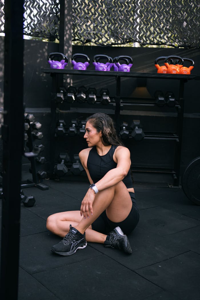 Woman in sportswear sitting on gym floor surrounded by kettlebells and dumbbells post-workout.